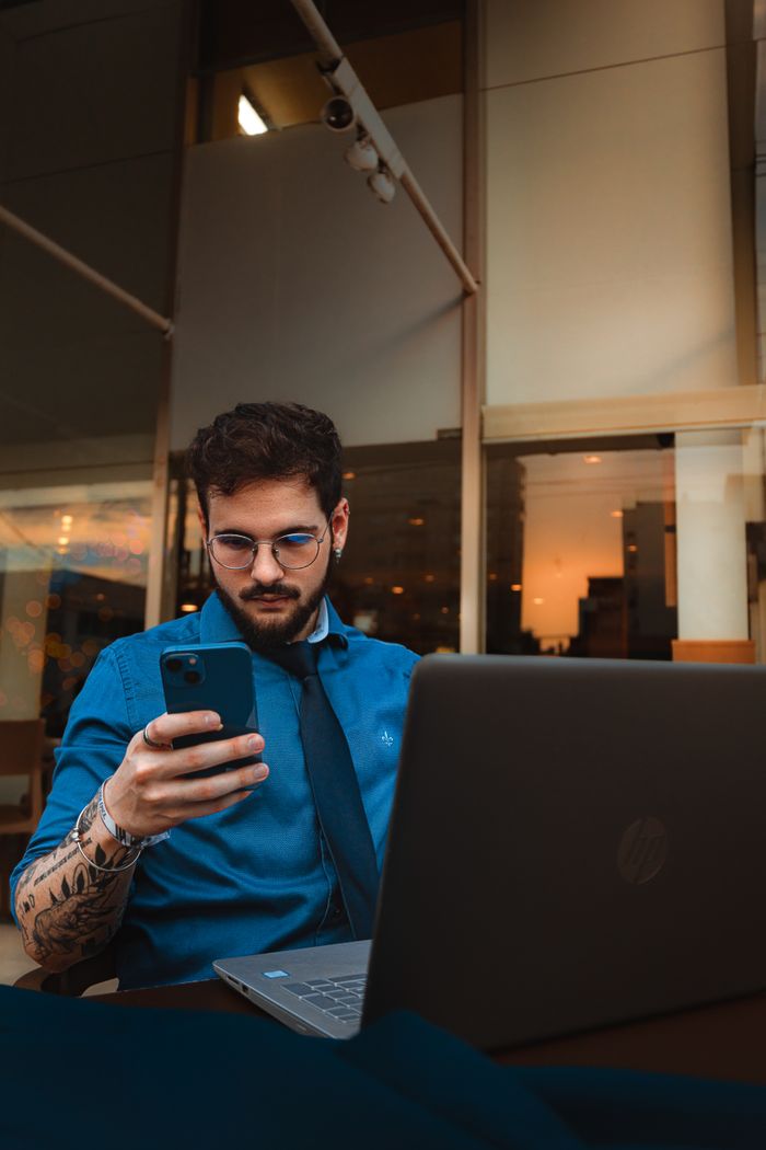 A man with a beard and eyeglasses focuses on his smartphone, sitting indoors with a laptop.